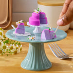 Miniature purple cake with white flowers on a green stand, with a hand holding a small fork.