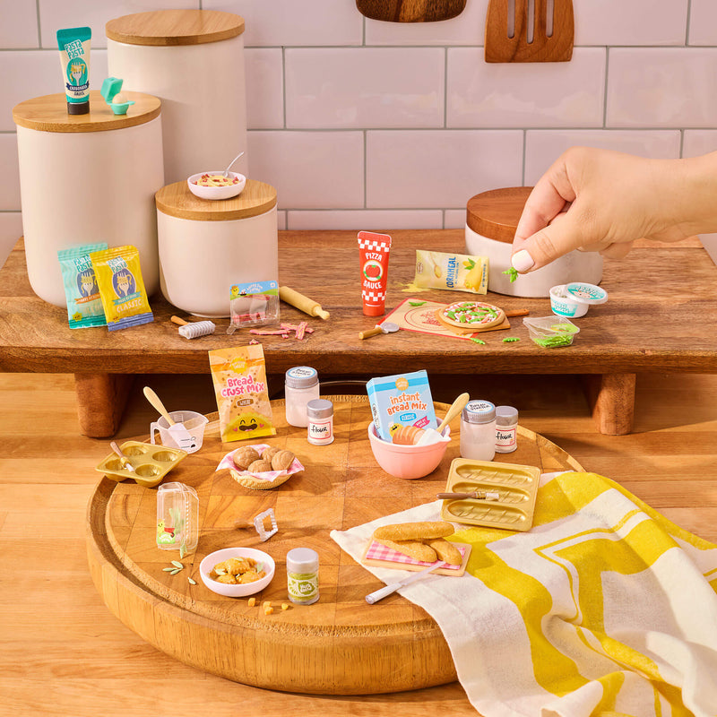 Miniature food items and kitchen accessories on a wooden surface with a hand interacting with them.