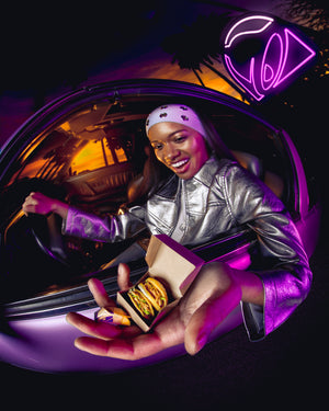 Person in a car holding Miniverse Taco Bell tacos with a neon Taco Bell logo sign in the background