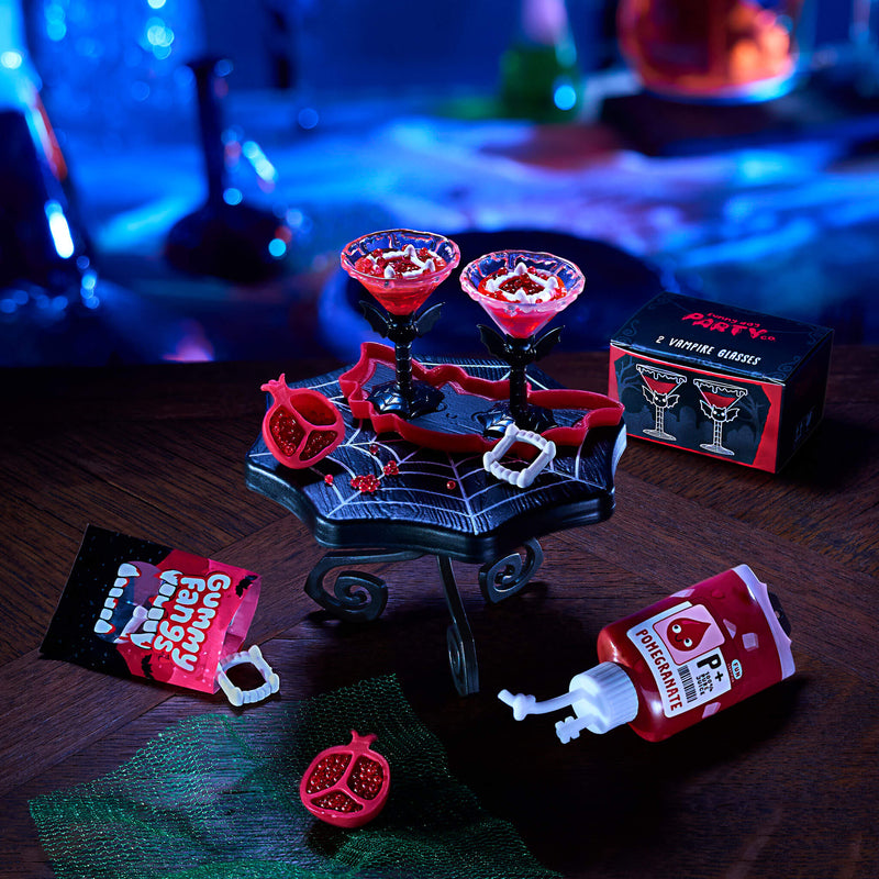 Halloween-themed cocktail set with two red cocktail glasses on a black spiderweb stand, plus fake blood, a pomegranate prop, a squeeze bottle, and packaging on a dark tabletop with a blue-lit blurred background.