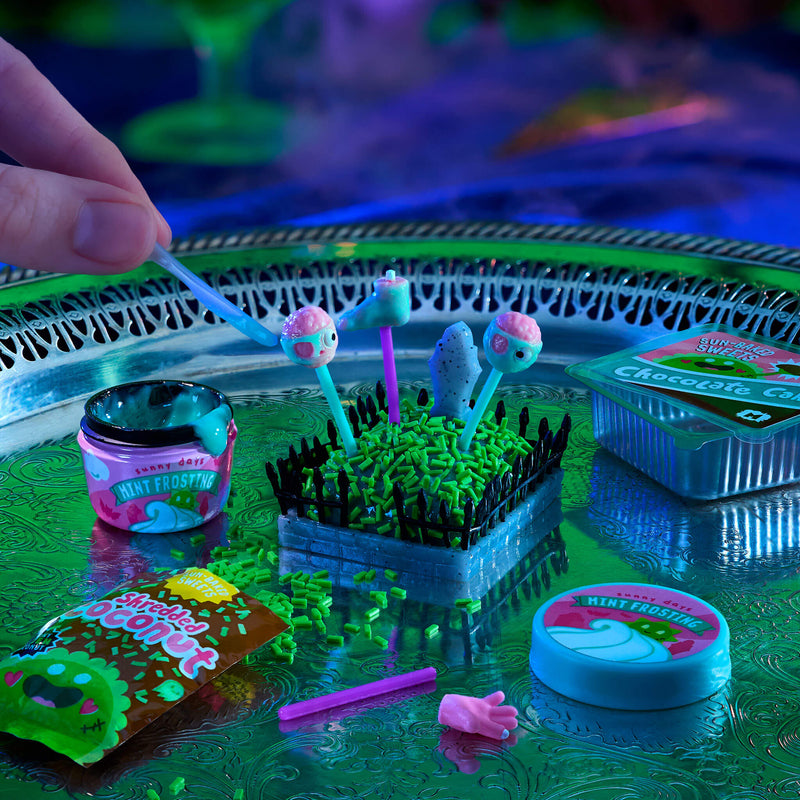 A hand is placing a mint-frosted pick into a small plastic garden display on a reflective green tray, surrounded by other mint-frosted candy and accessories under blacklight.
