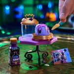 A miniature Halloween-themed setup on a reflective green table, featuring a purple and blue decorated cupcake on a small purple stand, a clear jar with a gold lid, a small bottle labeled 'Syrup,' a colorful 'Gummy Bug' bag, and a hand holding a small spatula.