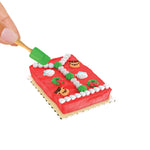 Red frosted cake with decorations and a hand holding a mini green spatula on a white background
