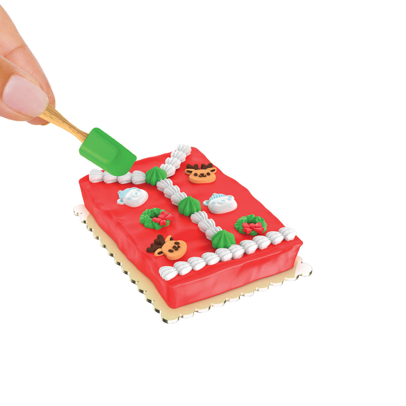 Red frosted cake with decorations and a hand holding a mini green spatula on a white background