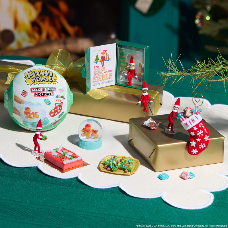 Various mini-Elf on the Shelf holiday items on a white table runner with a green table cloth underneath.