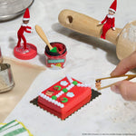 Red frosted cake with decorations and a hand holding a mini reindeer with mini tweezers to put on the cake. Two Elf’s in the kitchen setting background.