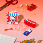 Mini notebook with Hello Kitty on the front, a small red box, with a pencil sharpener and scissors in the back on a peach background.