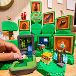 A Minecraft-themed display set up on a wooden table, featuring green blocky trees, framed pixel-art panels, small character figures, a cow, a pig, a flying pink creature, and a hand holding one of the figures. Visible branding includes Minecraft and a prominent Miniverse.