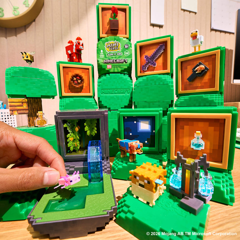 A Minecraft-themed display set up on a wooden table, featuring green blocky trees, framed pixel-art panels, small character figures, a cow, a pig, a flying pink creature, and a hand holding one of the figures. Visible branding includes Minecraft and a prominent Miniverse.