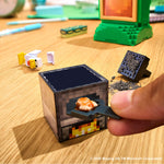 A close-up of a hand placing food into a small pixel-style cube stove toy on a wooden tabletop. The stove has a dark top, pixelated gray sides, and a yellow-orange flame graphic on the front.