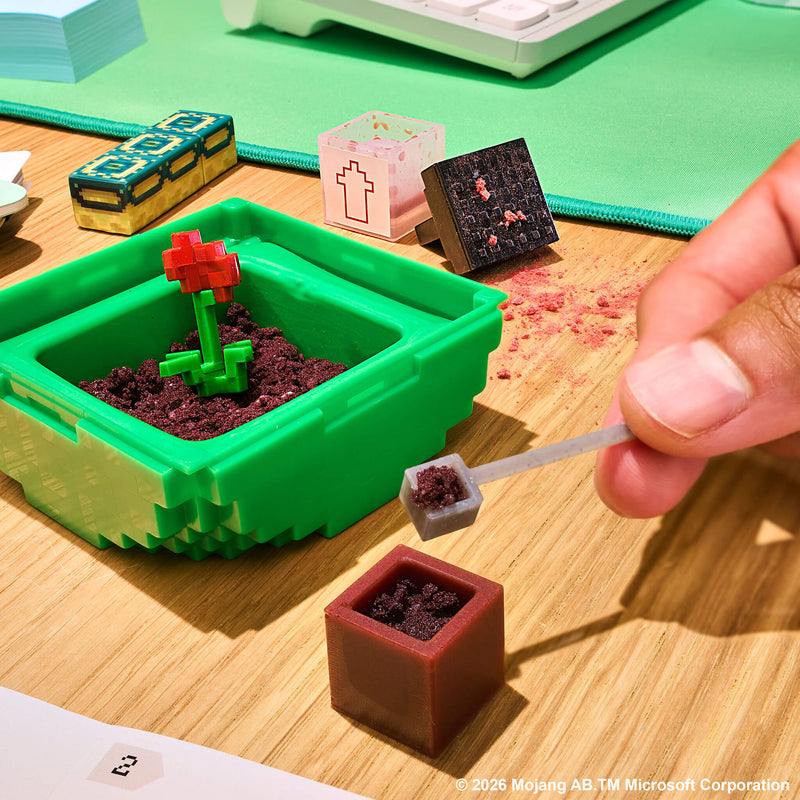 A close-up of a hand placing dirt into a miniature brown pot on a wooden desk. The scene includes a bright green planter with dark soil and a small red flower, along with other tiny  gardening-like pieces and a green desk mat in the background.