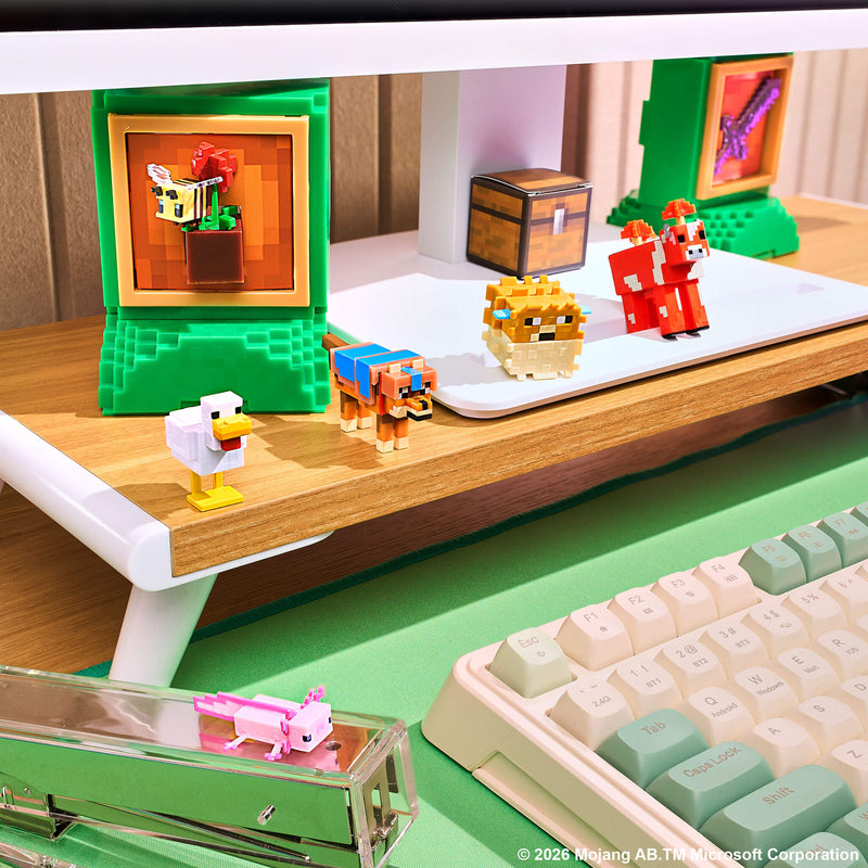 A close-up of a desk setup featuring small blocky figurines inspired by Minecraft, including a chicken, a dog, a sheep, and a cow, arranged on a white monitor arm and desk shelf alongside a keyboard and monitor.