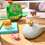 Minecraft Minis set displayed on a wooden desk with a green packaging, small pixelated animal figures, a book, and a mouse in a styled room setting.