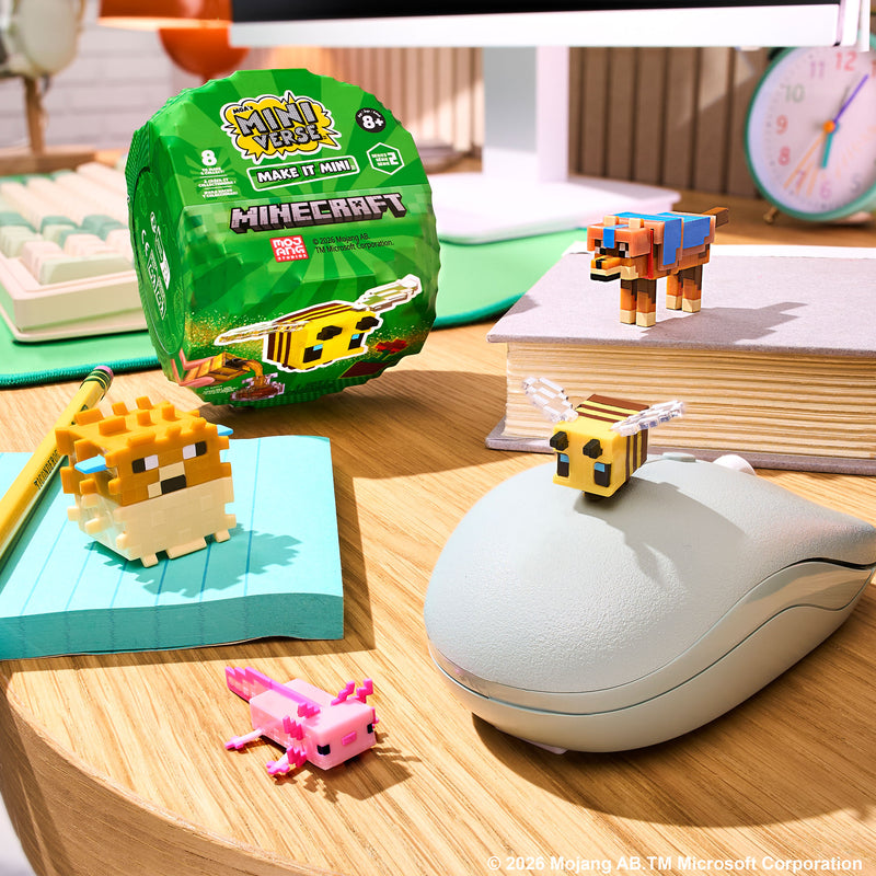 Minecraft Minis set displayed on a wooden desk with a green packaging, small pixelated animal figures, a book, and a mouse in a styled room setting.