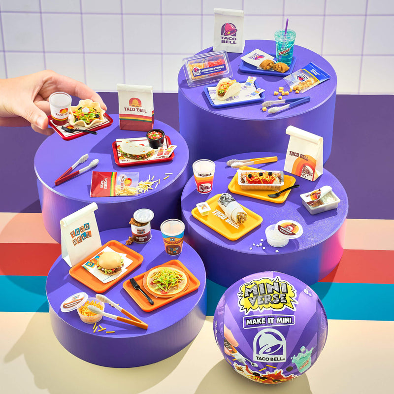 Miniature Taco Bell food sets sitting on purple, round, platforms with a hand interacting , against a white tiled wall.