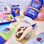 A hand squeezes a purple bottle of Taco Bell Sour Cream onto a taco, with other Taco Bell Flatbread and condiment packaging arranged around it on a tabletop.