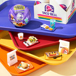 A bright, colorful arrangement of stacked cafeteria trays and Taco Bell-branded items, photographed from above at a slight angle. The scene uses bold purple, red, orange and yellow trays layered vertically.