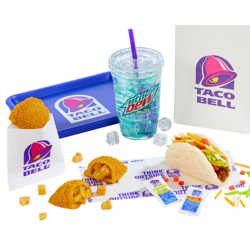 Taco Bell menu items and branding arranged on a white background.  A clear plastic cup with a purple straw containing a turquoise-blue drink labeled “Mtn Dew Baja Blast.” Blue Taco Bell tray with the Taco Bell bell logo printed on it. White paper bags showing the purple-and-pink Taco Bell bell logo. Fried, Apple Empanada (one cut open to show orange filling). A folded soft taco with seasoned filling, shredded cheese and diced red/green toppings.