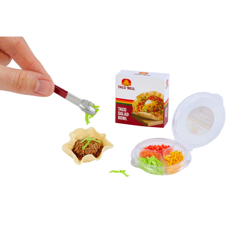 A miniature plastic taco salad bowl set shown on a white background, with a hand using a small pick to serve food from the tiny bowl. The set includes a small taco salad bowl, a small box labeled Taco Bell, and an open round container with compartments filled with colorful pretend salad ingredients.