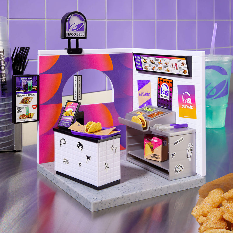 A small drive-thru / counter fixture with the Taco Bell bell logo mounted on top. Purple, pink and orange branded panels and posters, including a vertical “LIVE MÁS” poster. Food items on the counter: two tacos on paper and a condiment/salad station with diced toppings sitting on a silver table top with purple tiled background.