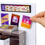 A Taco Bell ordering/condiment area with branded cards on the white, brick wall and a small menu display at the top. A hand is adding a card to the wall.