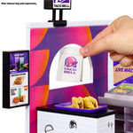 A Taco Bell-themed playset or counter scene showing a hand placing a small paper takeout bag through a service window with a purple and orange patterned panel.