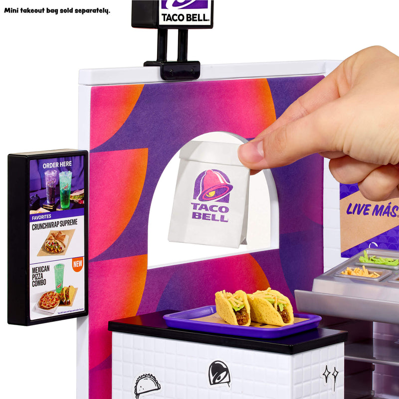 A Taco Bell-themed playset or counter scene showing a hand placing a small paper takeout bag through a service window with a purple and orange patterned panel.