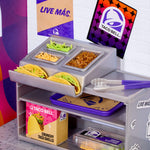 A miniature toy Taco Bell food display stand with mini tacos, small food containers, tongs, and branded packaging on a tabletop. The display includes Taco Bell signage and a colorful branded box.