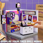 Taco Bell counter scene set against purple tiled walls. The service counter, food prep area, and a hand placing a tiny red tray on the countertop. Mini food bins containing taco fillings, a drink dispenser, one is labeled “ICE CUBES.” Text on the bottom “ Display Your Taco Bell Minis!”