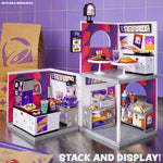 A miniature restaurant set with two stacked levels, featuring purple and pink Taco Bell themed walls, serving tables with food, drink cups, a counter, and accessories, shown with a brown paper bag and text "Stack and Display" on a purple-tiled background.