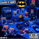 A Batman-themed mini toy and collectible display arranged on a blue grid surface, including a bat-shaped car, comic books, a Bat-light prop, and a purple Batman-themed  case.