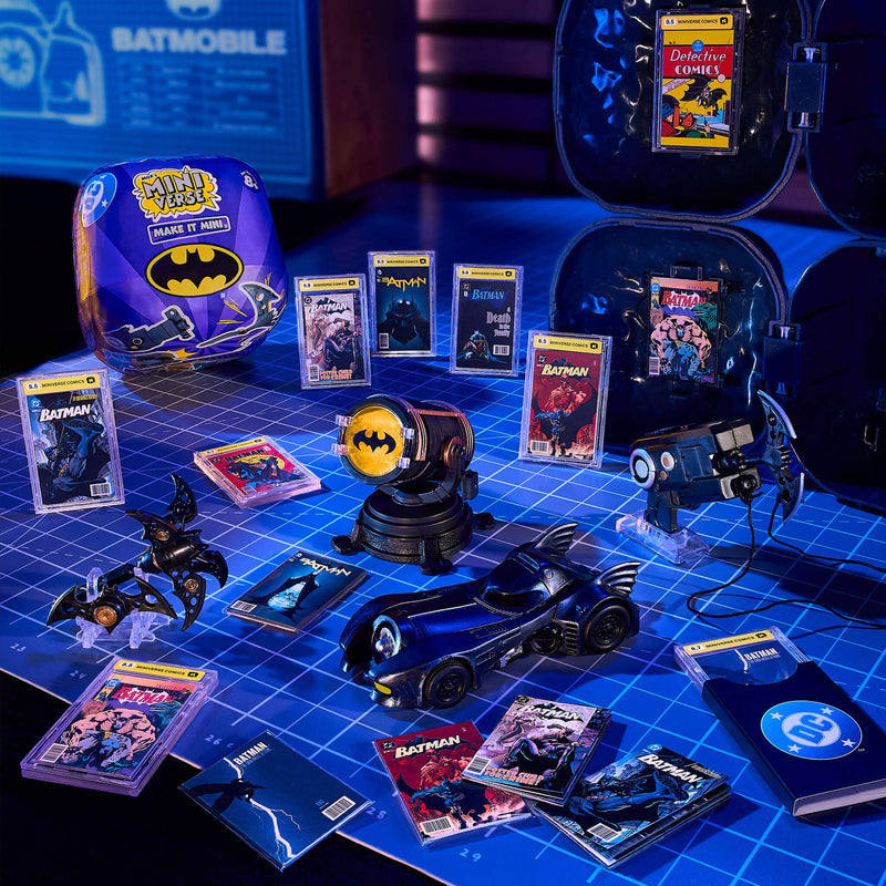 A Batman-themed mini toy and collectible display arranged on a blue grid surface, including a bat-shaped car, comic books, a Bat-light prop, and a purple Batman-themed  case. 