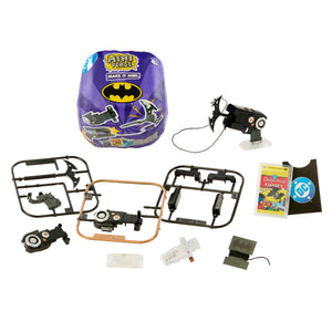 A Batman-themed toy set laid out on a white background, including a purple packaged round item, a small black flying 'bat' with a wire, several black plastic parts on frames, a small book,  and a few small accessories.