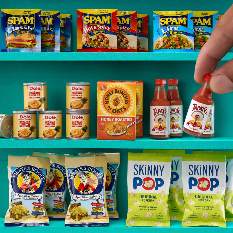 Three teal shelves with mini food; top has different packages of Spam, the middle had Dole Pineapple, Honey Bunches of Oat, jar of sauce; the bottom has Pirates Booty and Skinny Pop popcorn bags.