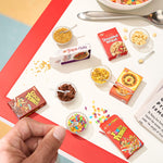 Mini cereal boxes on a red and white table, with a hand holding a spoon over a cereal bowl.