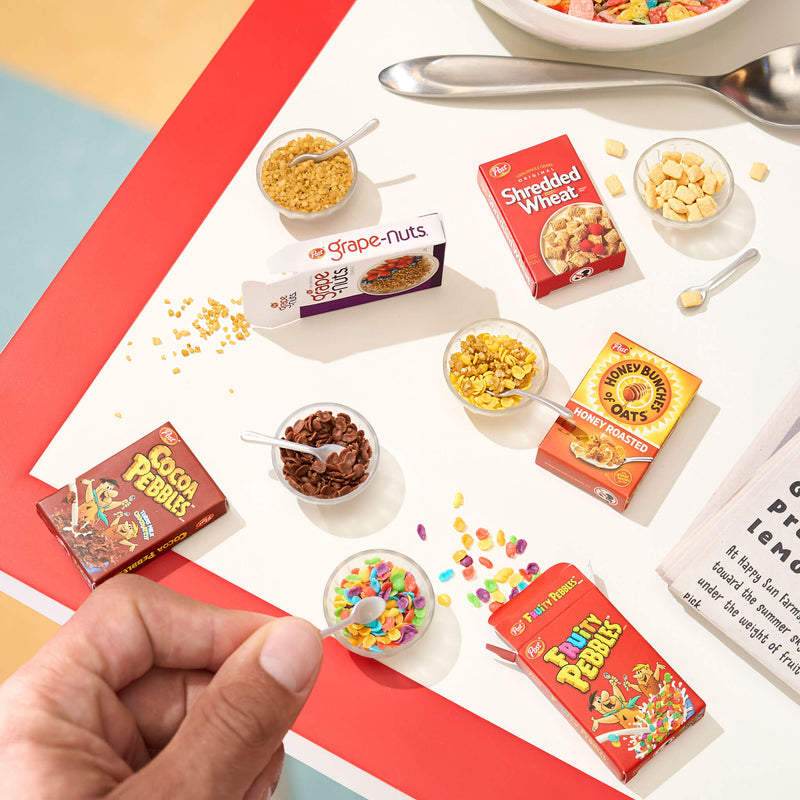 Mini cereal boxes on a red and white table, with a hand holding a spoon over a cereal bowl.