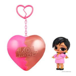 Small doll with short black hair wearing a pink dress standing next to a heart shaped keychain on a white background