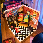 Miniature record store display with vinyl records, guitar, and amplifier on a brown table with blue curtains in the background.