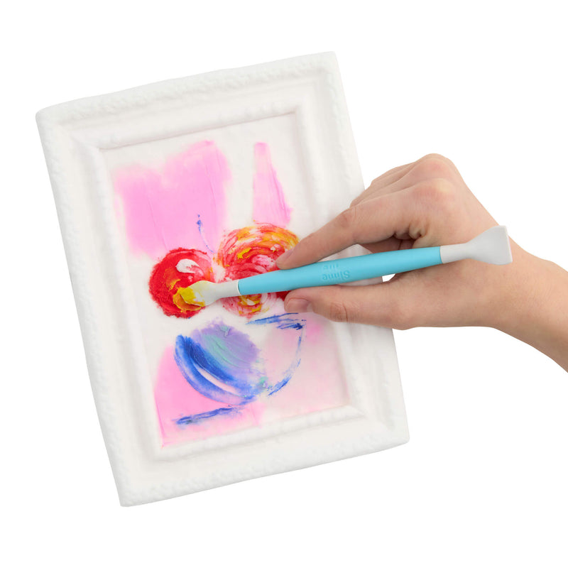 A hand holding a light blue paint brush , painting on a small white textured square canvas that features abstract pink, red, yellow, and blue brushstrokes, set against a plain white background.