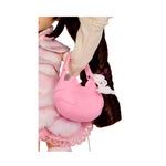 A pink cat-shaped handbag with a white charm, shown being carried by a doll wearing a pink and white outfit against a white background.