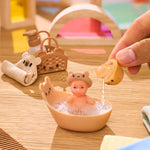 Mini toy baby in a bathtub with a hand pouring water, surrounded by bath accessories on a wooden surface.