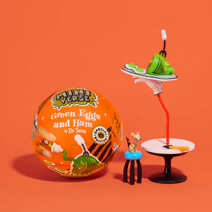 A 'Green Eggs and Ham' surprise toy set on a solid orange background, featuring a orange ball with printed graphics, a small figurine of a mouse on a stool, and a miniature tray with a hand holding a green ham.