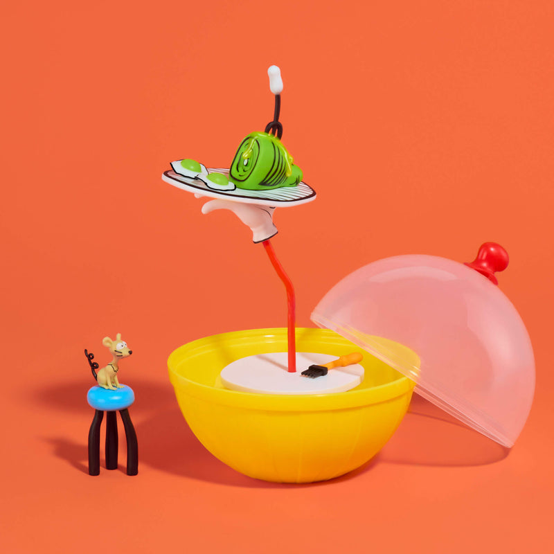 A toy set on a solid orange background, featuring half of a yellow ball packaging with a hand stand, a translucent dome lid,  a small figurine, and a miniature plate with a green ham item.