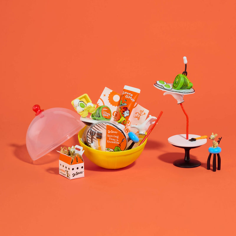 A miniature toy set featuring a yellow bowl filled with small food items, a translucent dome lid, a tiny carton, and a small table with a serving stand, all arranged on a solid orange background.