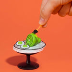 A miniature green ham and two mini eggs on a small illustrated plate, with a human hand holding a tiny orange brush above it against a solid orange background.