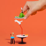 A miniature tabletop scene with a human hand holding a small tray with green ham and eggs above a white table, hand stand, and tiny mouse figurine, set against a solid orange background.