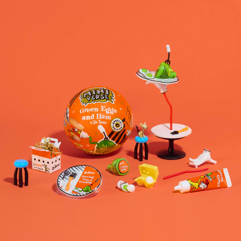 A miniature tabletop set of Dr. Seuss-themed items on a solid orange background, including a round orange 'Green Eggs and Ham' ball, a small white-and-black table, a green egg, a tube of 'Green Eggs' cream, a plate, and other tiny illustrated accessories.