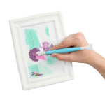 A hand holding a light blue and white paint brush inside a white frame, showing a light green and purple painted design.