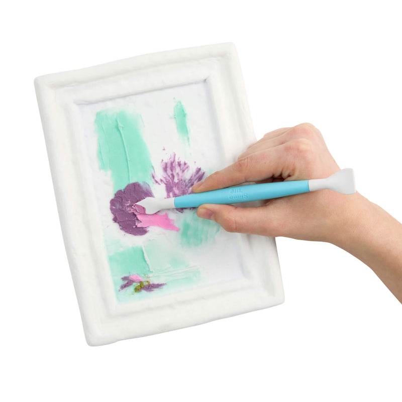 A hand holding a light blue and white paint brush inside a white frame, showing a light green and purple painted design.
