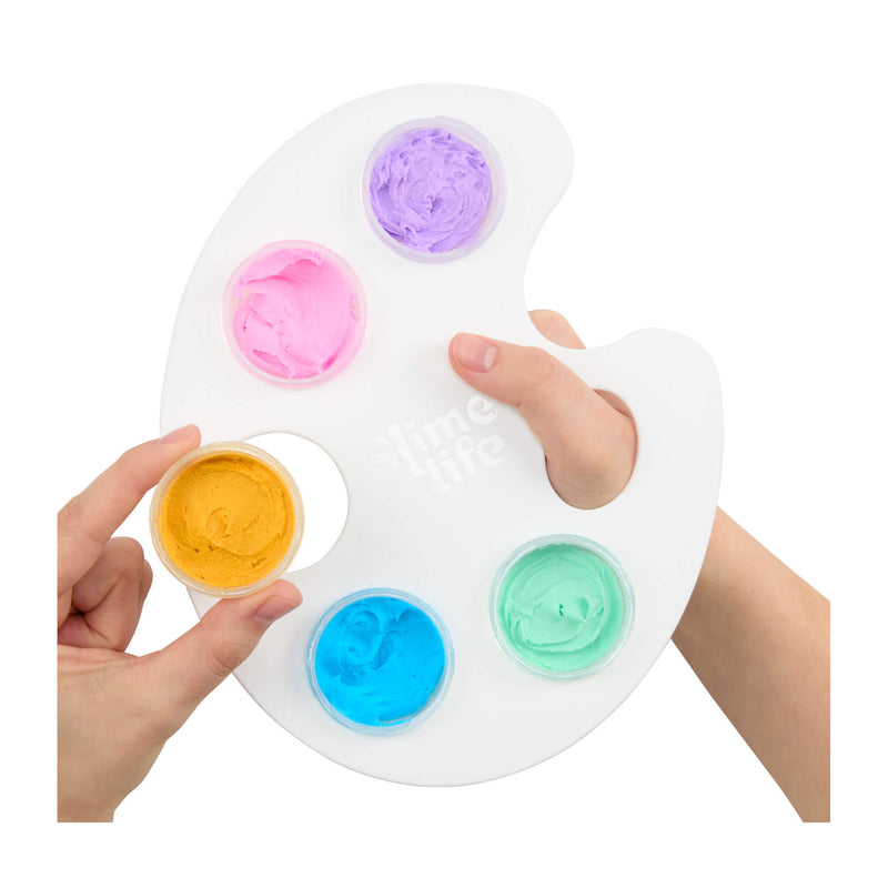 A white artist palette with five circular wells holding pastel purple, pink, yellow, blue, and mint green paints, shown with two hands against a white background.